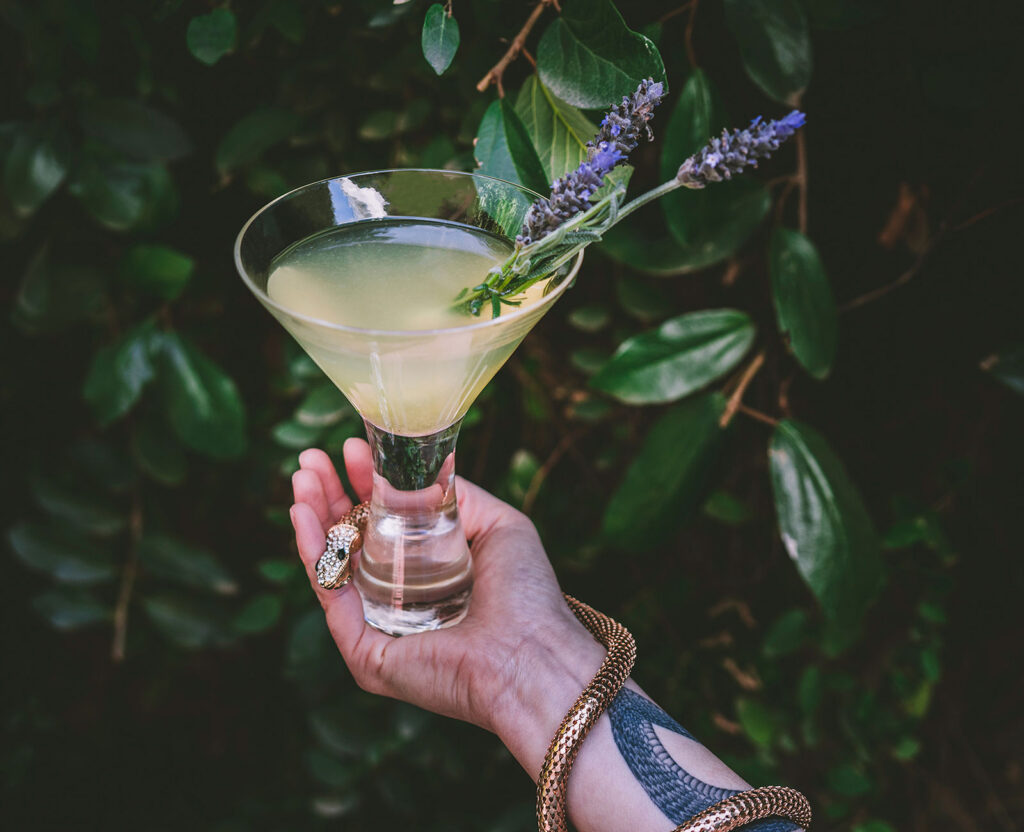 Lavender Twist Gin Cocktail held by a hand with a gold serpent and serpent tattoo