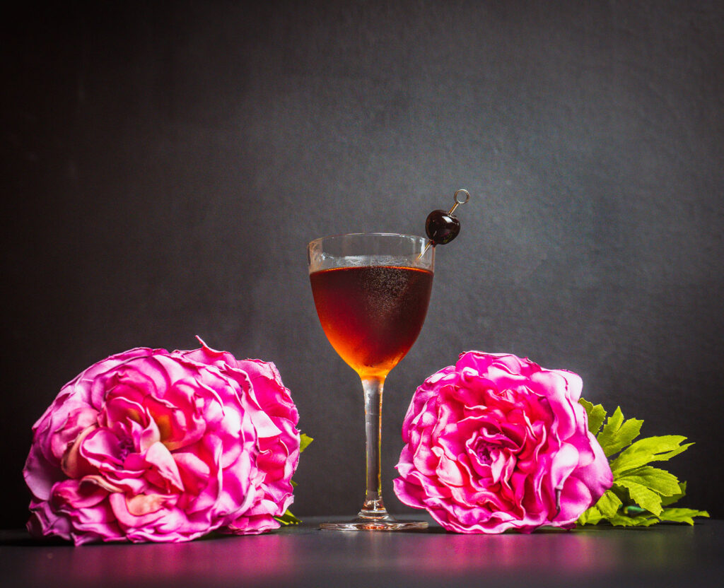 Bijou cocktail with a cherry garnish and two pink flowers on the sides